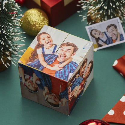 Alt text: A festive gift box with a photo print of a smiling man and woman wrapped around it, placed among holiday decorations, with a snow-frosted tree and gold ornament visible.