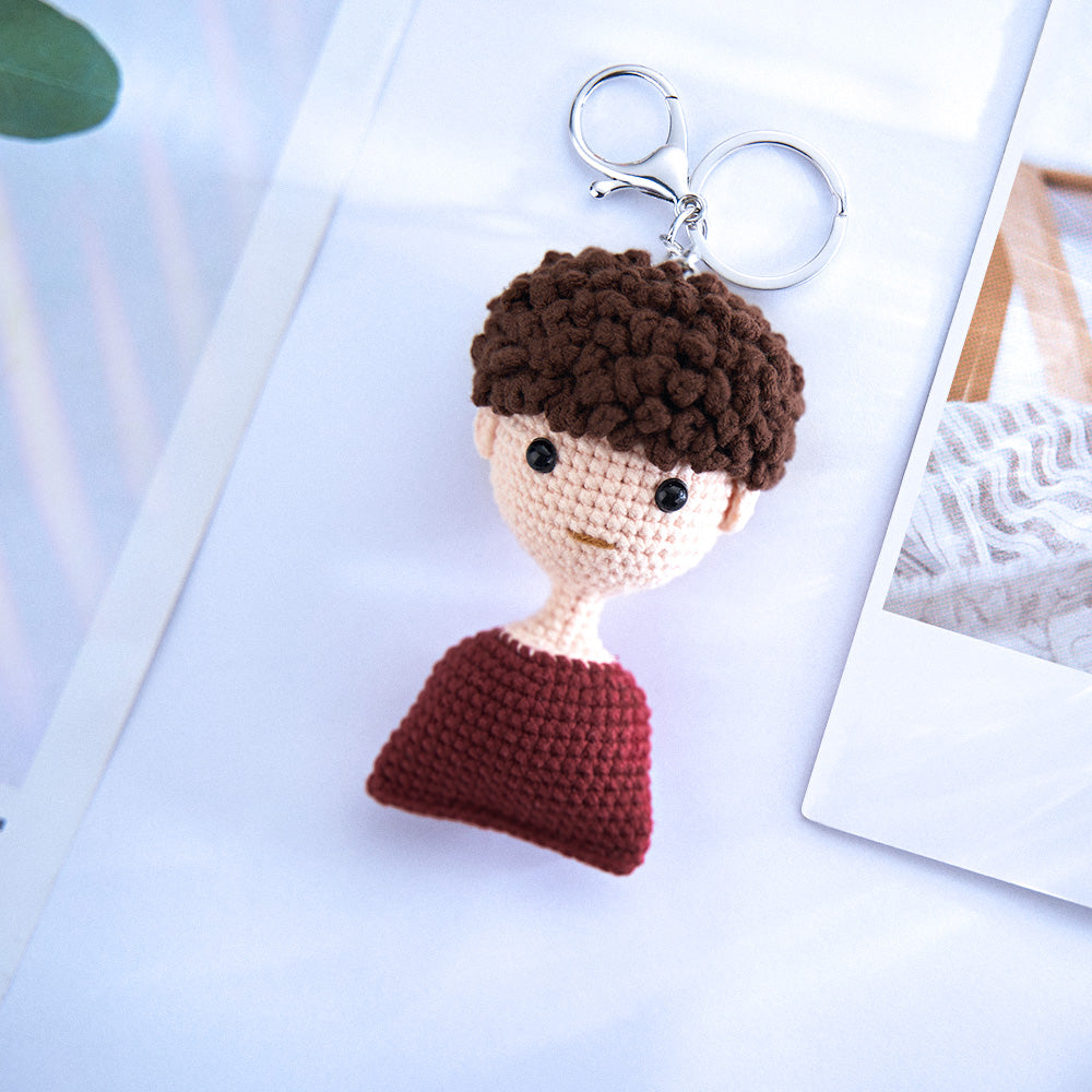 Crocheted keychain in the shape of a person with a brown curly hair, black eyes, and a red top, resting on a white surface with soft background.