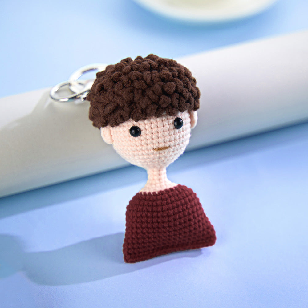 A handmade crochet keychain in the shape of a character's head and shoulders with curly brown hair, black eyes, and a red shirt, set against a light blue background.