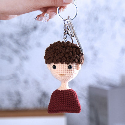 Hand holding a crocheted keychain with a stylized human figure featuring a brown hair, a simple face, and a red shirt, against a soft-focus background.