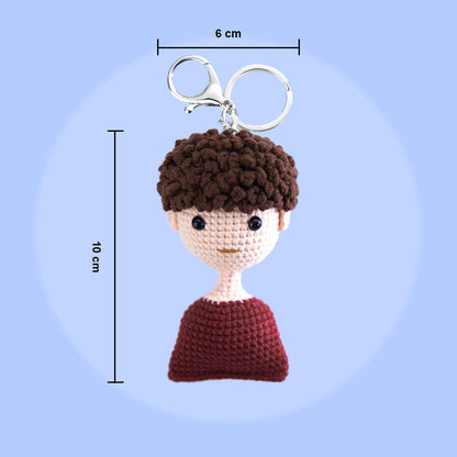 A crocheted keychain in the shape of a human head and upper torso measuring 10 cm in height, with a 6 cm keyring attached to the top, set against a blue background.