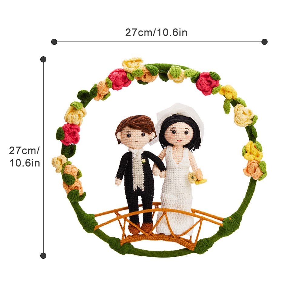 Crocheted bride and groom figurines standing on a bridge under a floral arch with dimensions labeled as 27 cm/10.6 in.