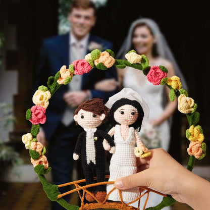 A person's hand holds crochet figurines of a bride and groom through a floral arch, with a blurred real-life couple in wedding attire in the background.