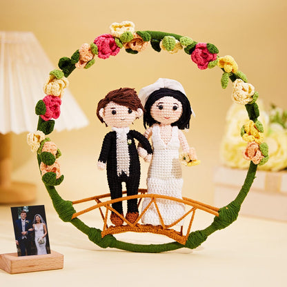 A crocheted representation of a bride and groom standing on a small bridge adorned with colorful flowers, with a real photo of a couple in the background.