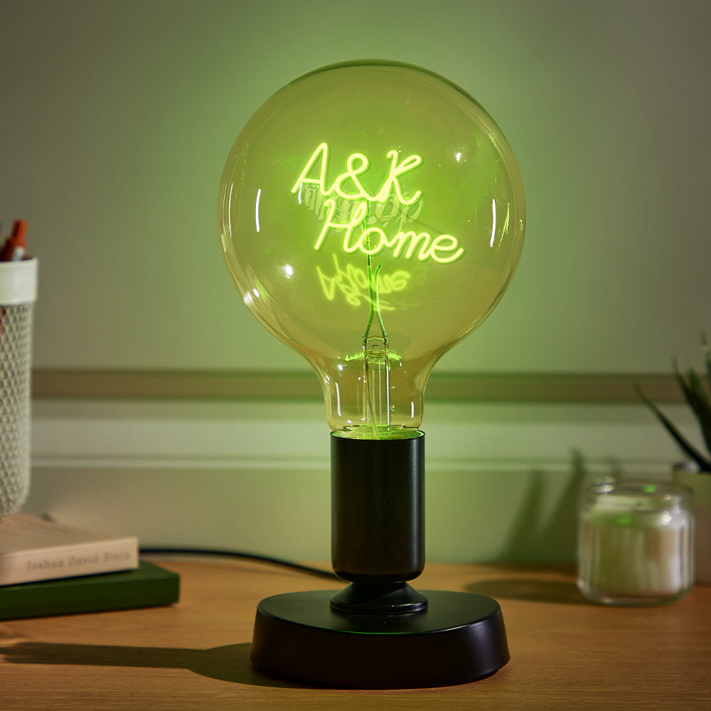 A large, glowing lightbulb on a dark base with "A&K Home" written inside it in neon script, placed on a wooden desk next to a book and a white pencil holder.