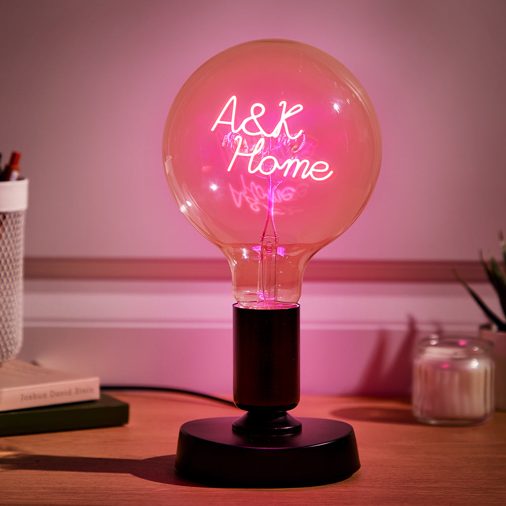 A neon sign within a large light bulb reads "Ask Me" glowing in pink on a desk with books and a candle, casting a warm light.