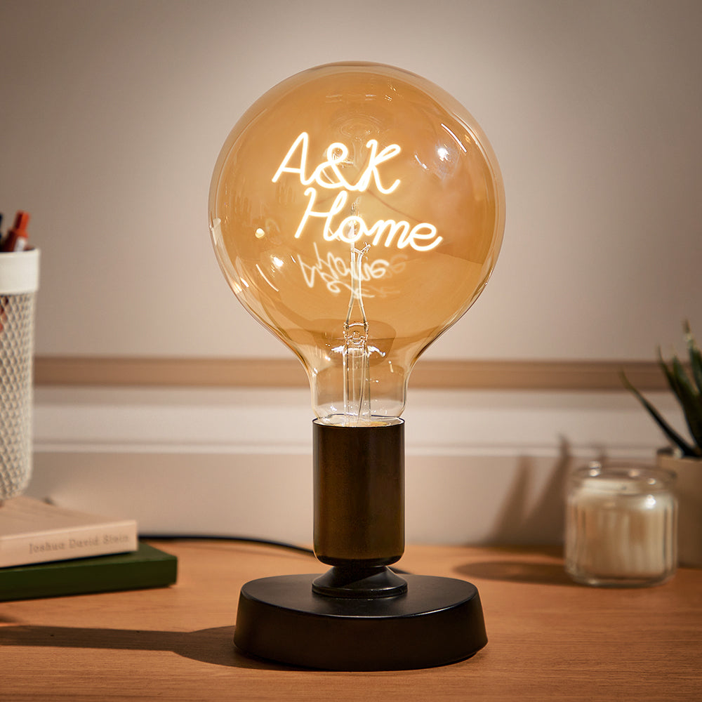 A large, illuminated round light bulb with "A&K Home" neon inscription on a brown lamp base, on a desk next to books and a pen holder.