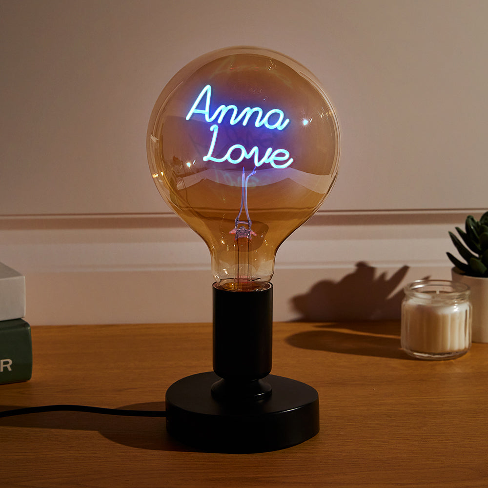 A lit custom neon sign within a large clear lightbulb that reads "Anna Love" on a table, with soft ambient lighting and a plant in the background.