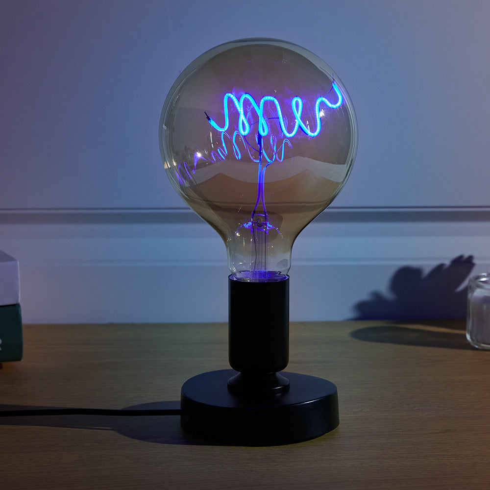 A plasma globe lamp on a desk emitting blue and purple tendrils of electricity inside a clear glass sphere, casting a soft glow in a dim room.