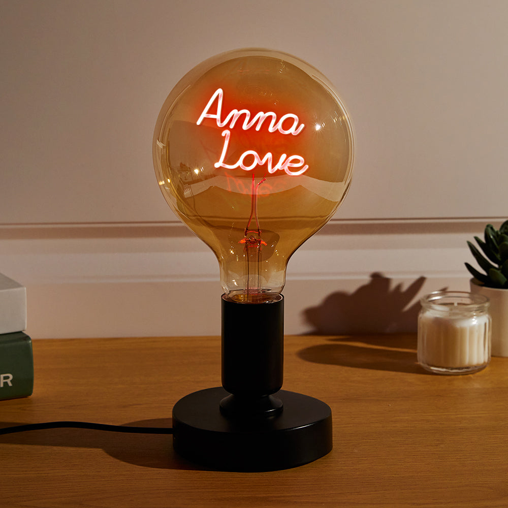 A large illuminated bulb on a black base with the red neon text "Anna Love" inside, on a wooden table next to a candle and plant.