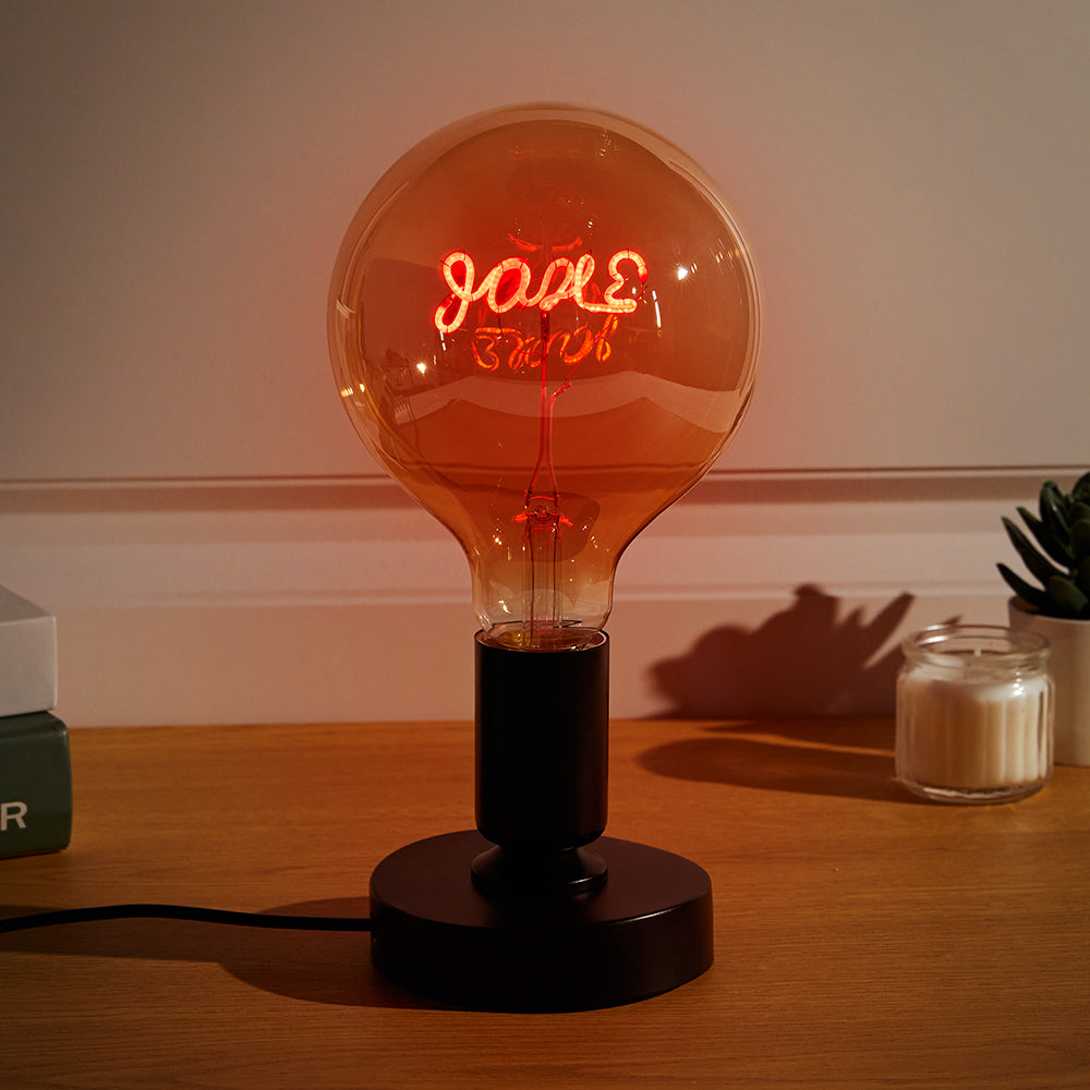 A glowing neon sign inside a large round lightbulb on a black base displays the cursive word "love," casting a soft red hue and shadow on the table.