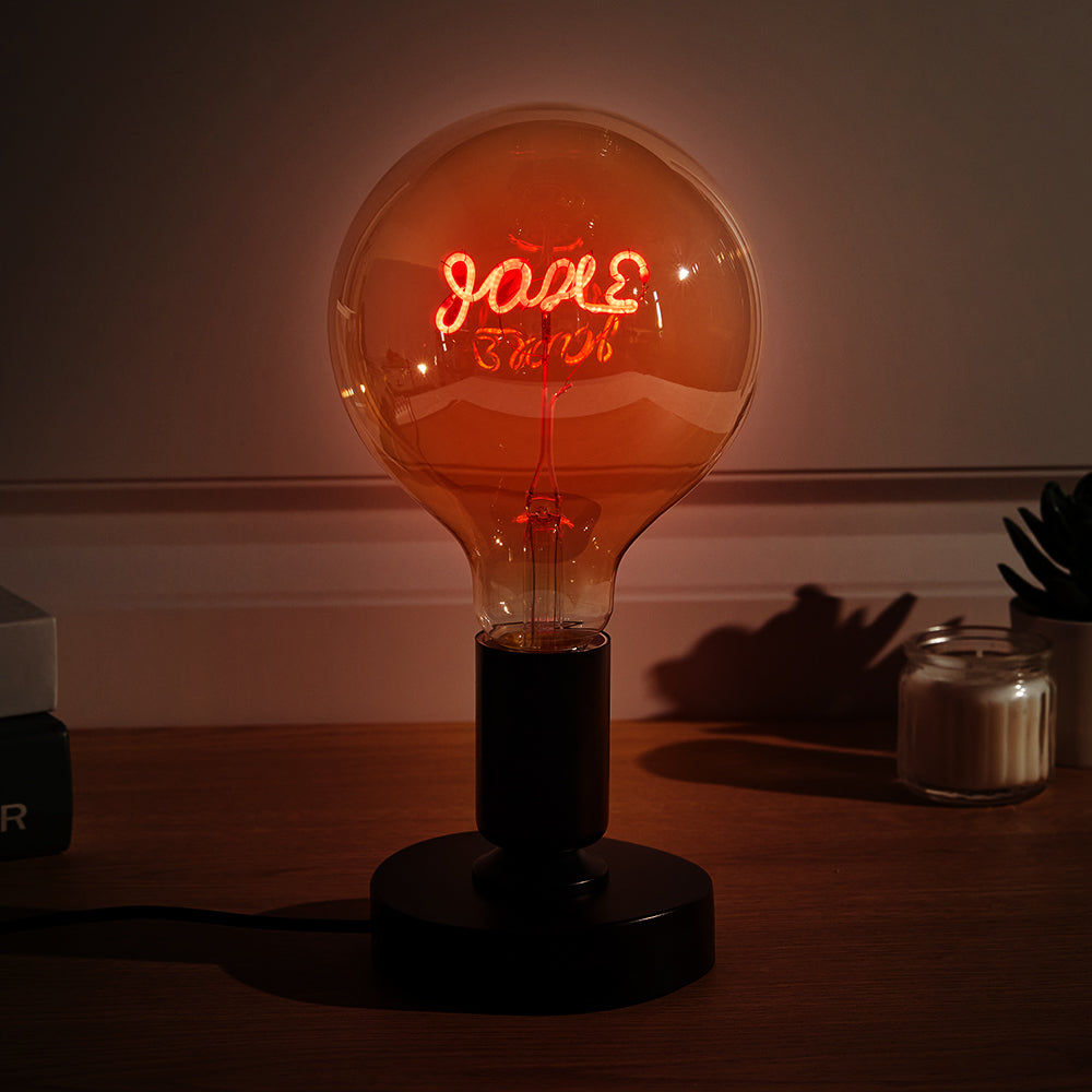 A large, round light bulb on a dark base emits a warm red glow with "love" written inside it, sitting on a wooden surface beside a small plant and candle.