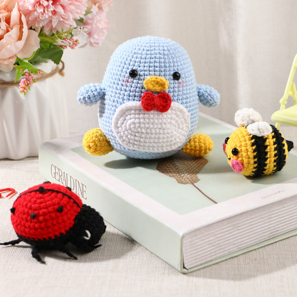 Handmade crochet toys including a blue penguin with a red bow tie, a bee, and a ladybug on top of a book titled "GERALDINE," with flowers in the background.