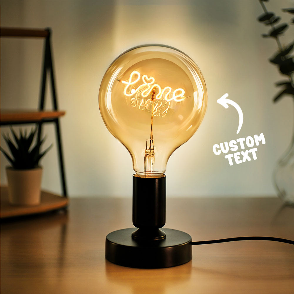 A decorative light bulb with the word "Love" illuminated in script on a black base, on a desk next to houseplants, with "CUSTOM TEXT" arrow indicator.