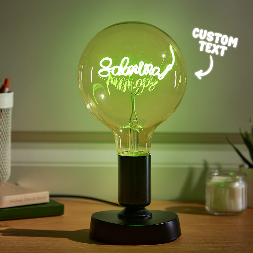 A large, illuminated glass light bulb with cursive script inside stands on a desk, with a "CUSTOM TEXT" neon sign in the background to the right.