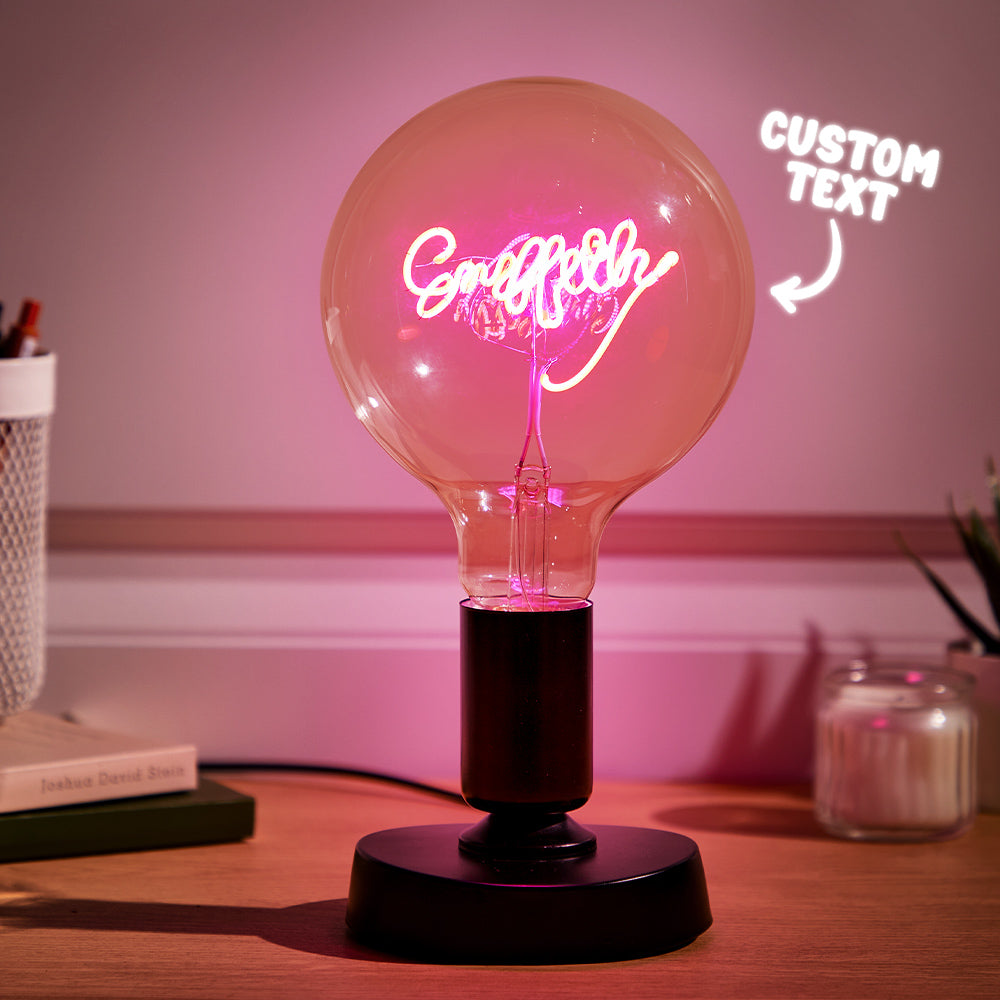 A neon sign with cursive text "Craftly" inside a large clear lightbulb on a black base, with a pink hue illuminating the room and "CUSTOM TEXT" in neon to the right.